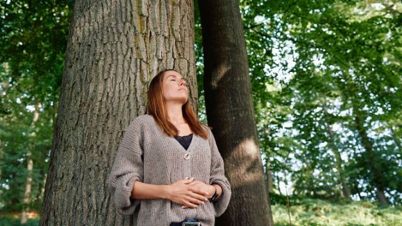 Junge Frau lehnt mit geschlossenen Augen an einem Baum im Wald und entspannt sich